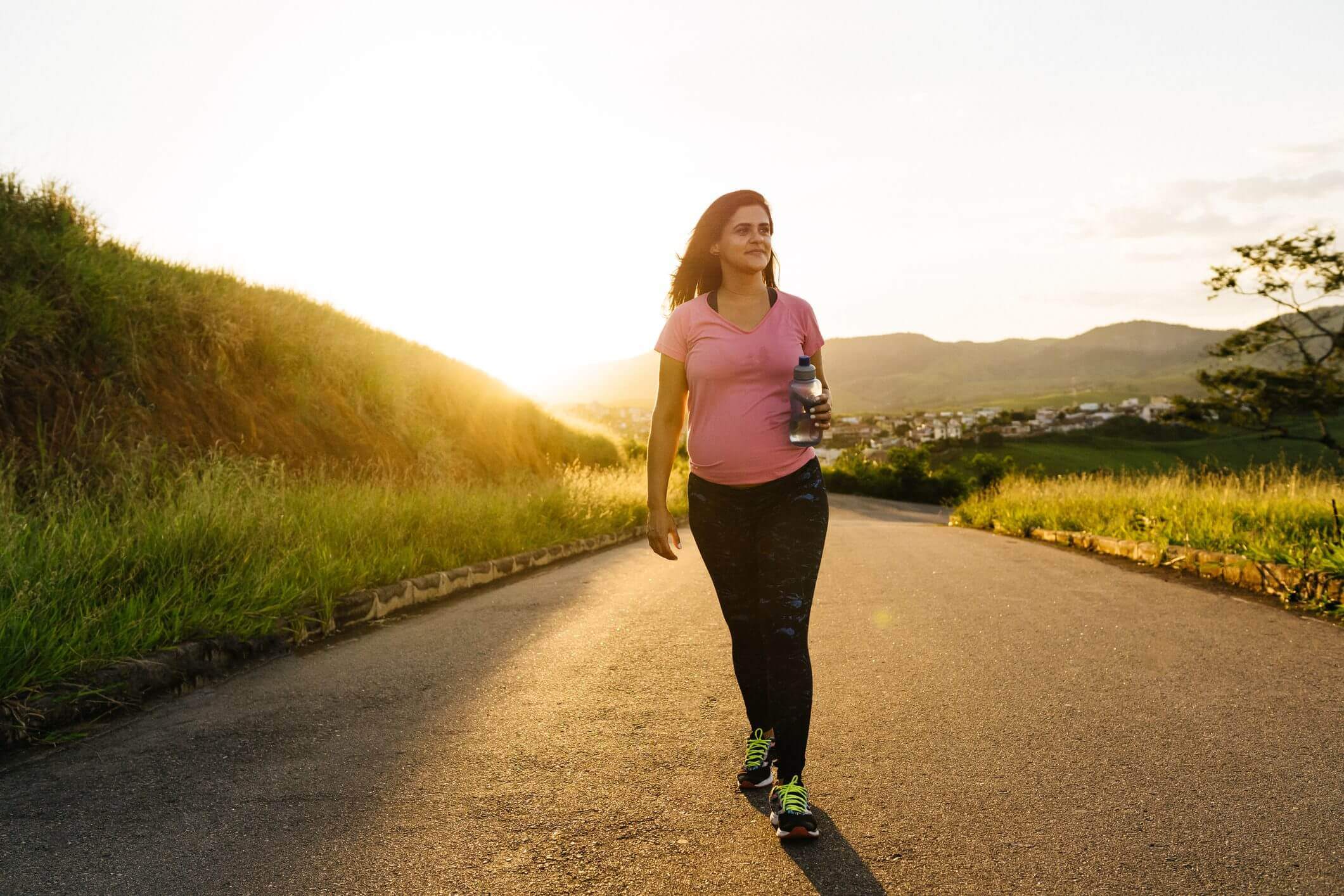 Woman walking on a sunlit road during sunset, holding a water bottle.
