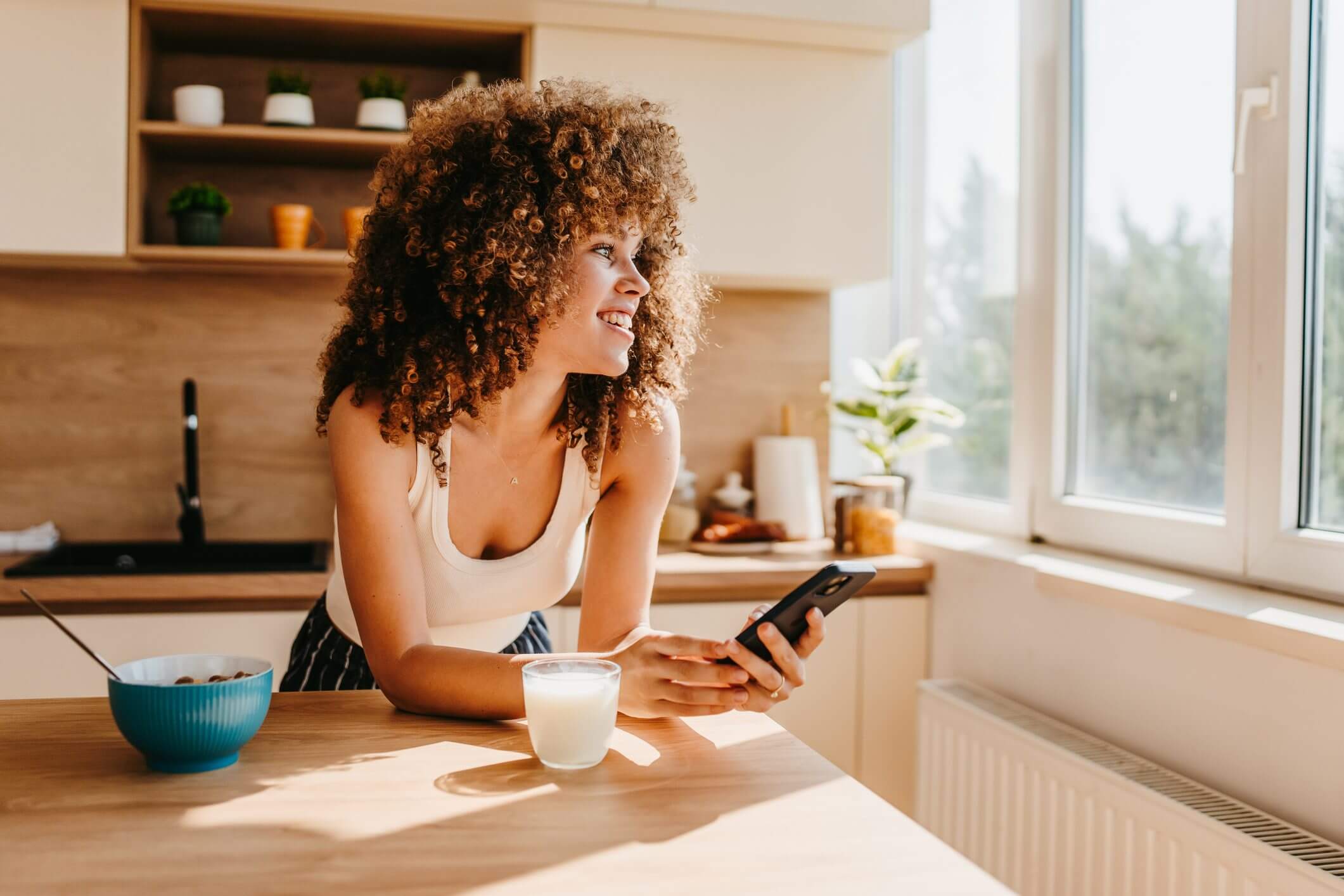 Woman with curly hair looking out window, holding phone and smiling.